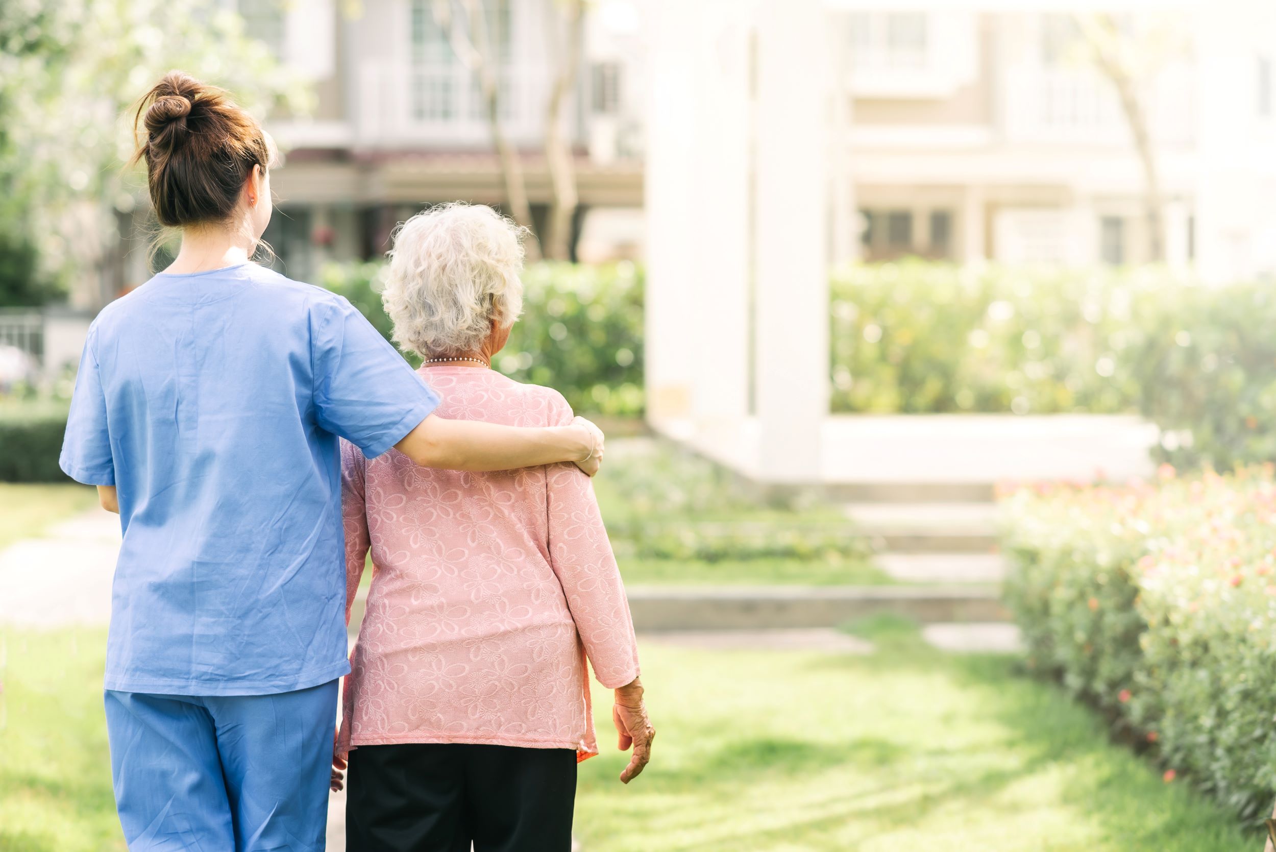A care worker and older woman facing away from the camera, walking outside towards a care home