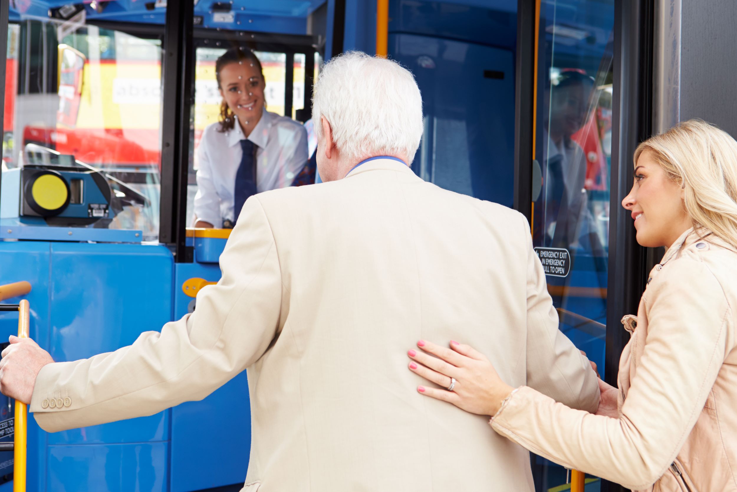 An older man and younger woman facing away, getting onto a bus. The bus driver is smiling