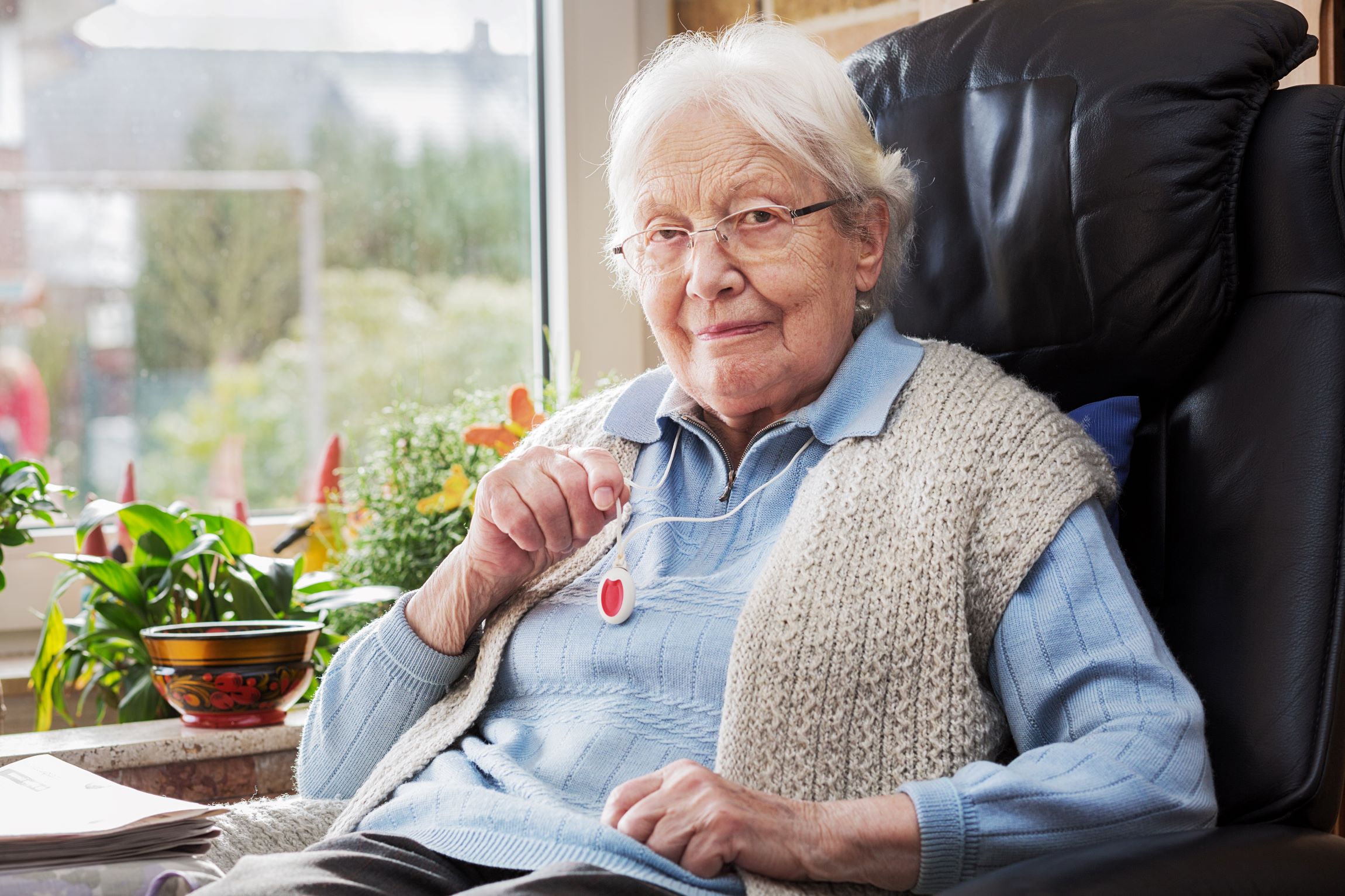 A woman looking at the camera is sat in an armchair, holding up her pendent alarm which she is wearing on her neck