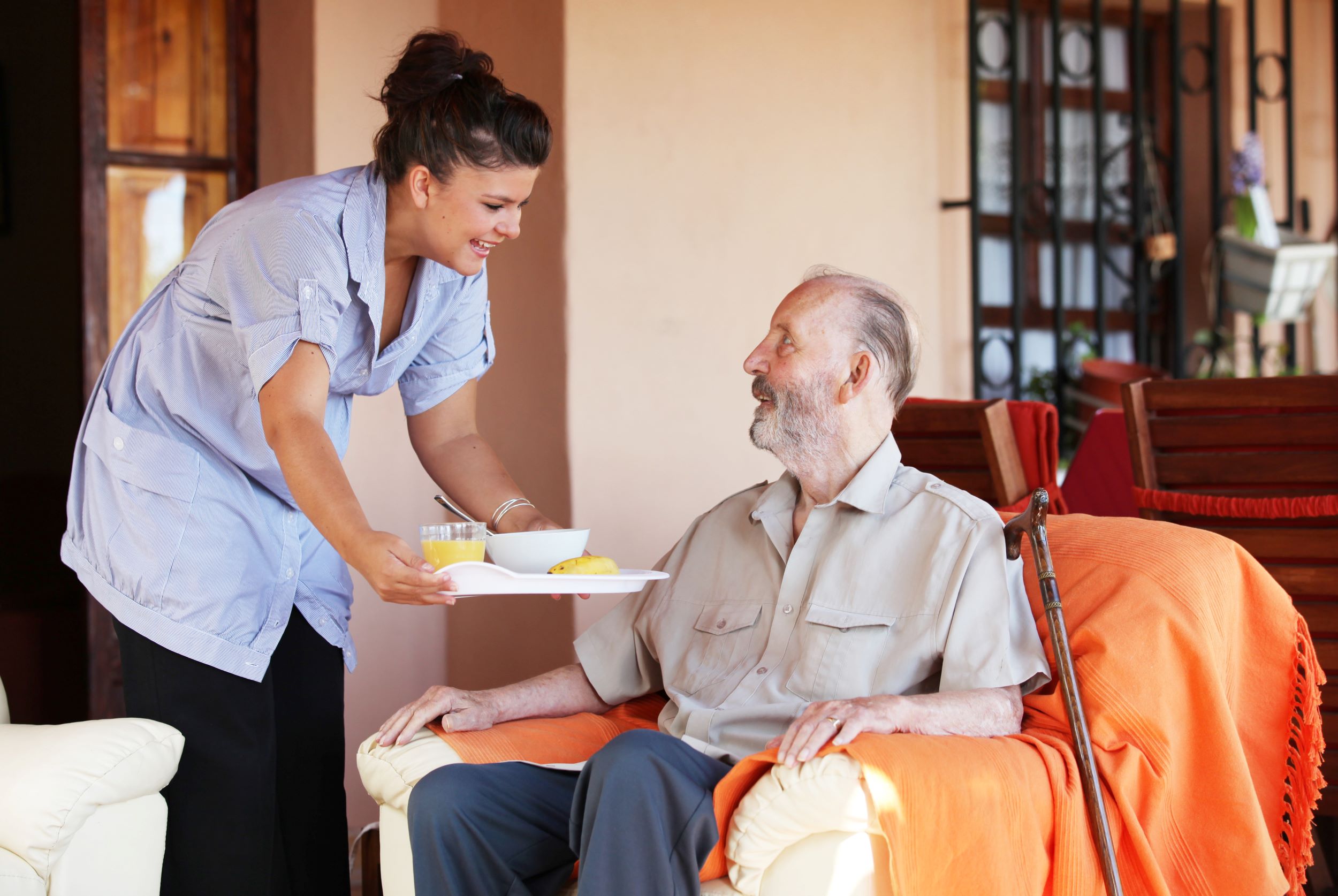 A care worker bringing lunch on a tray to a man sitting in an arm chair. He has a walking stick beside him