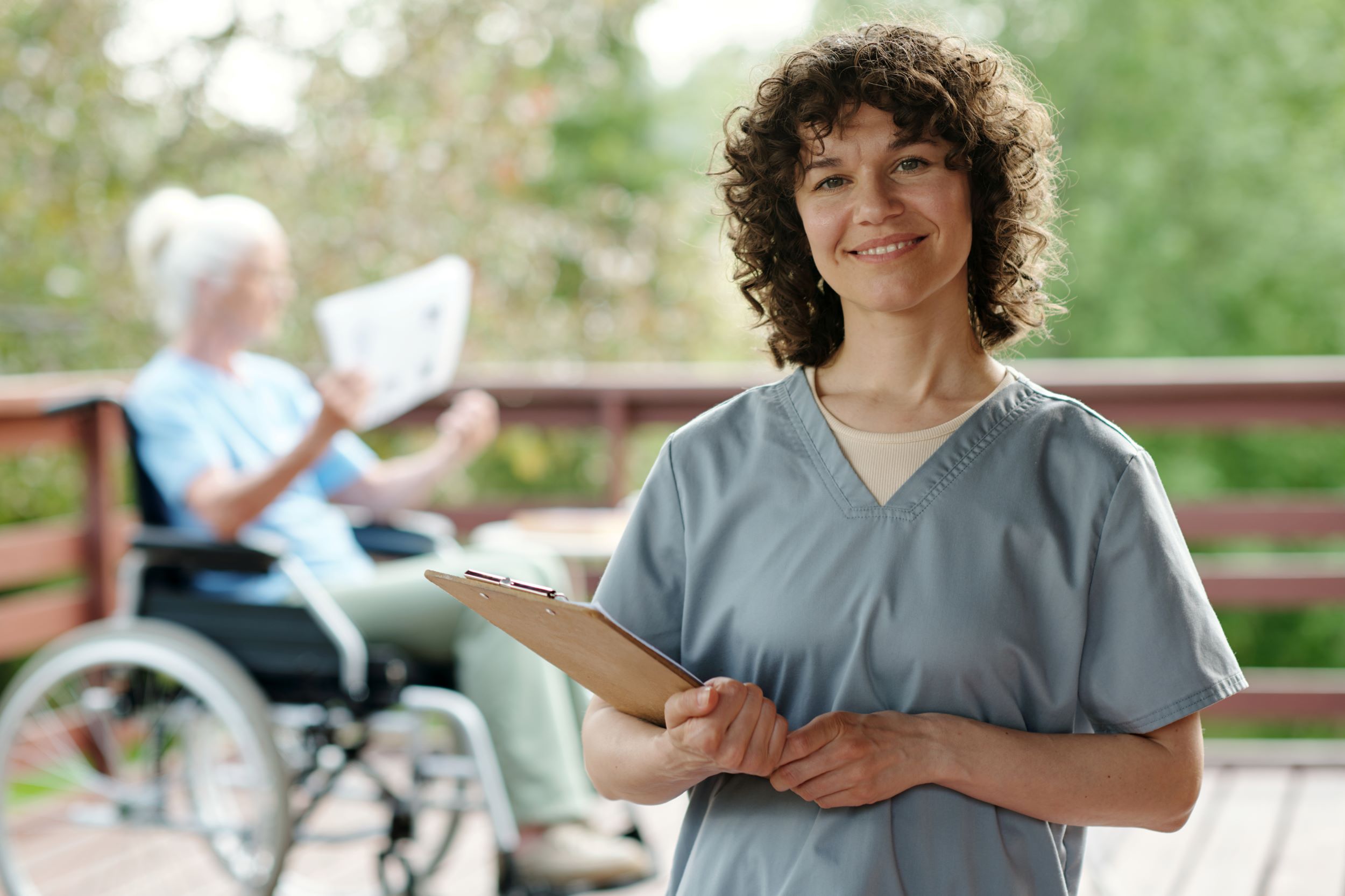 In the foreground is a care worker holding a clipboard. In the background is a woman in a wheelchair reading a newspaper