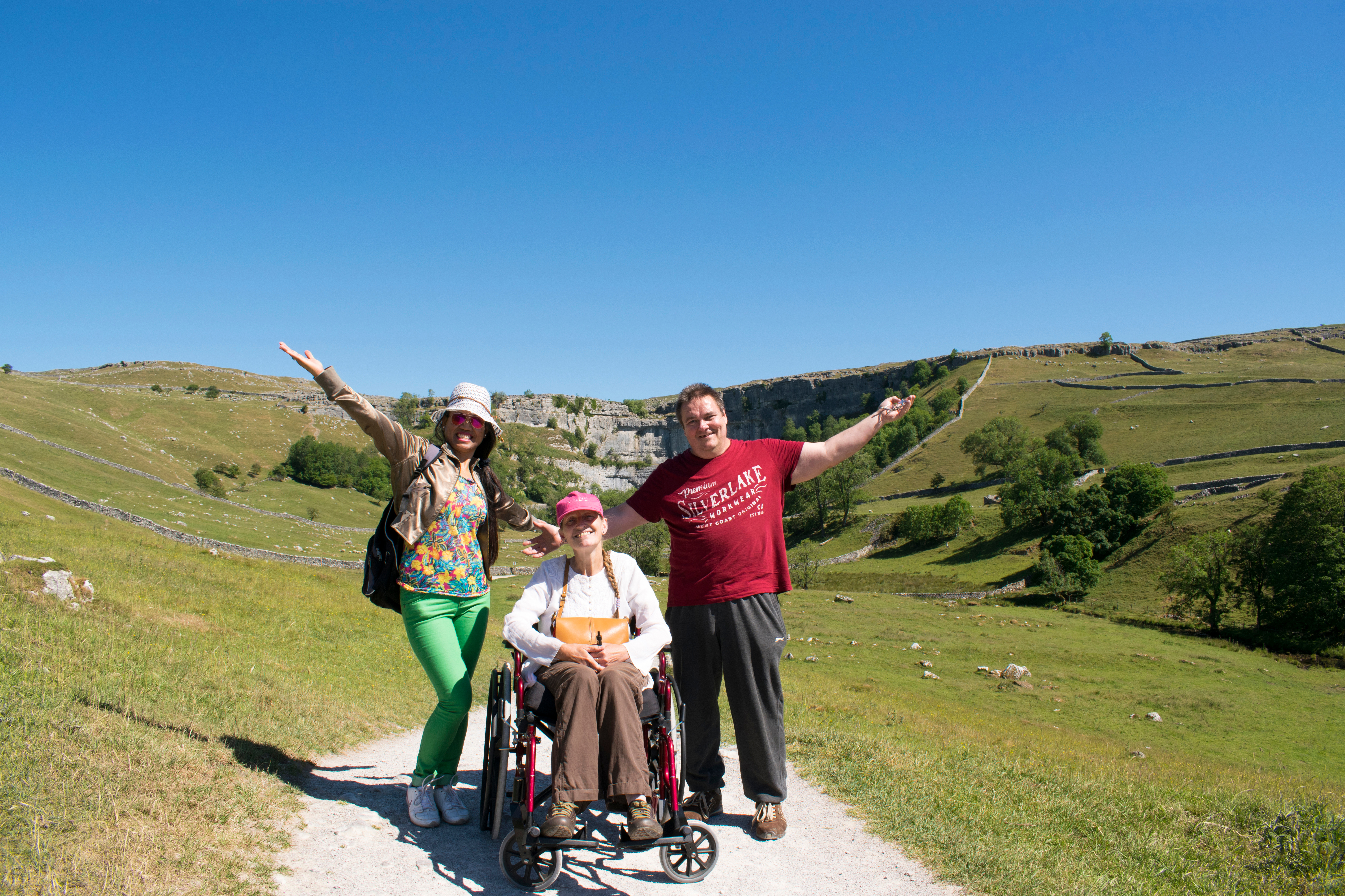 Three people outside on a hill top. Two people are stood either side of someone in a wheelchair, their arms are outstretched